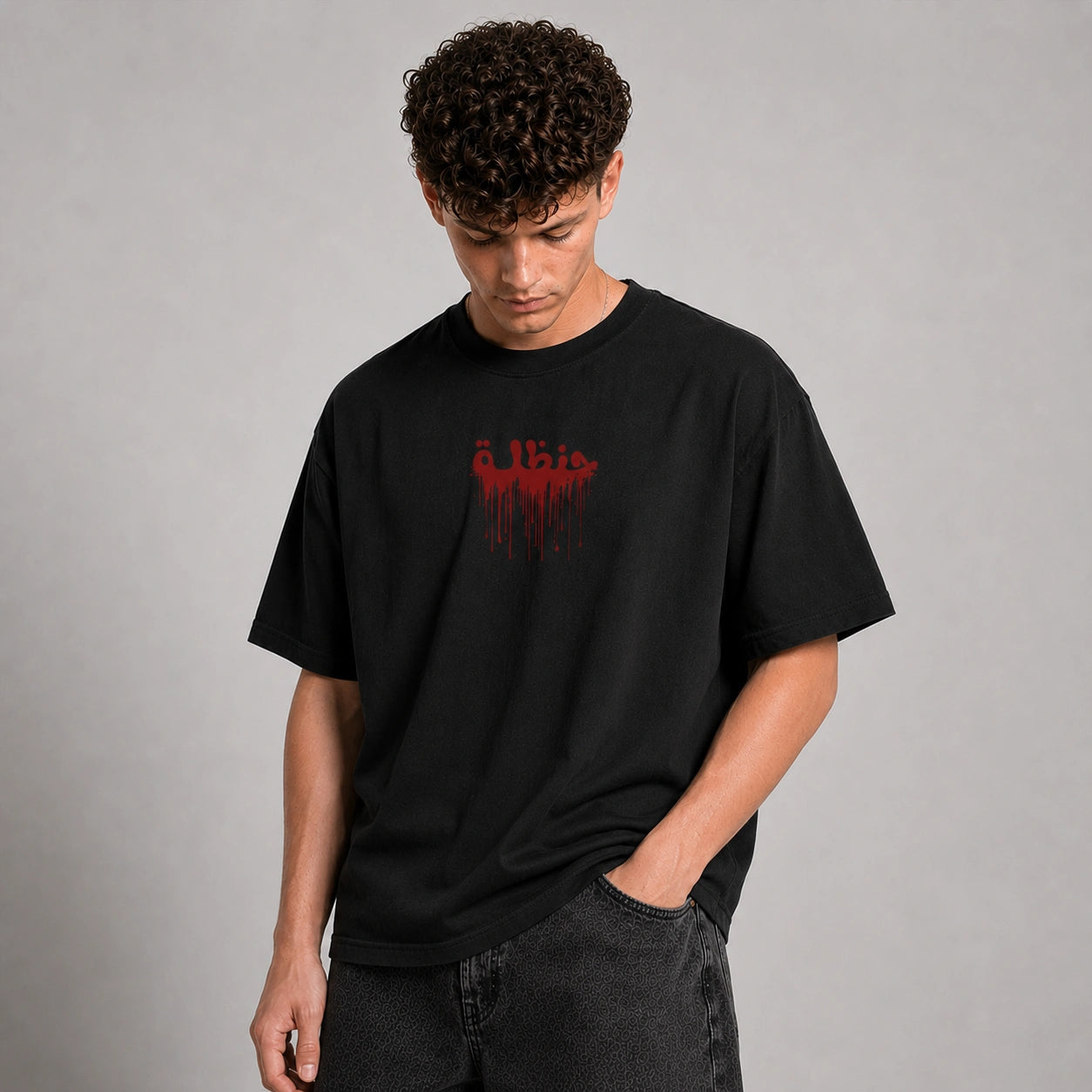 A man wearing a black crewneck t-shirt with a red dripping graphic text print on the chest, paired with dark gray jeans, standing against a neutral gray studio background.