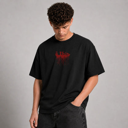A man wearing a black crewneck t-shirt with a red dripping graphic text print on the chest, paired with dark gray jeans, standing against a neutral gray studio background.