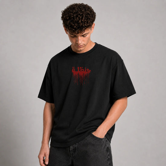 A man wearing a black crewneck t-shirt with a red dripping graphic text print on the chest, paired with dark gray jeans, standing against a neutral gray studio background.