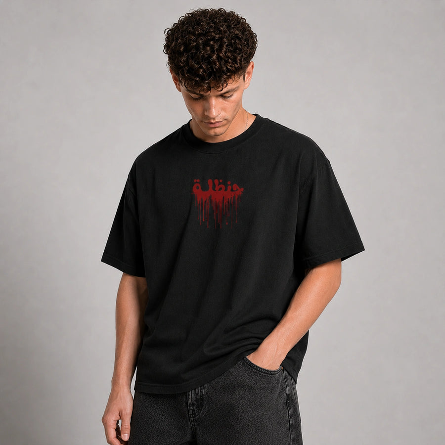 A man wearing a black crewneck t-shirt with a red dripping graphic text print on the chest, paired with dark gray jeans, standing against a neutral gray studio background.
