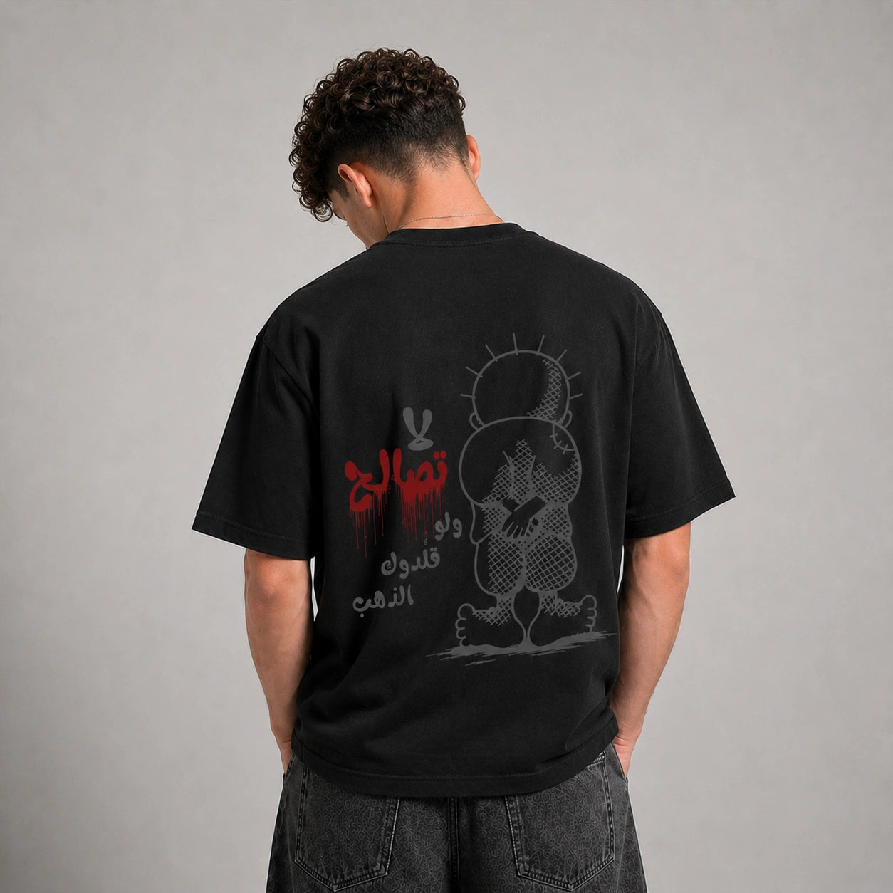 A man viewed from behind wearing a black graphic t-shirt with a red and gray design, paired with dark gray jeans, against a neutral gray studio background.