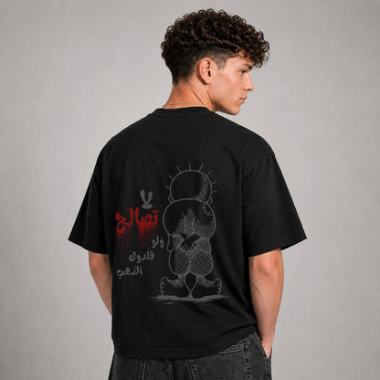 A man wearing a black t-shirt, shown from the back. The shirt features a graphic print of a cartoon character and Arabic text in red and gray.