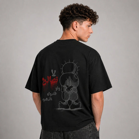 A man wearing a black t-shirt, shown from the back. The shirt features a graphic print of a cartoon character and Arabic text in red and gray.