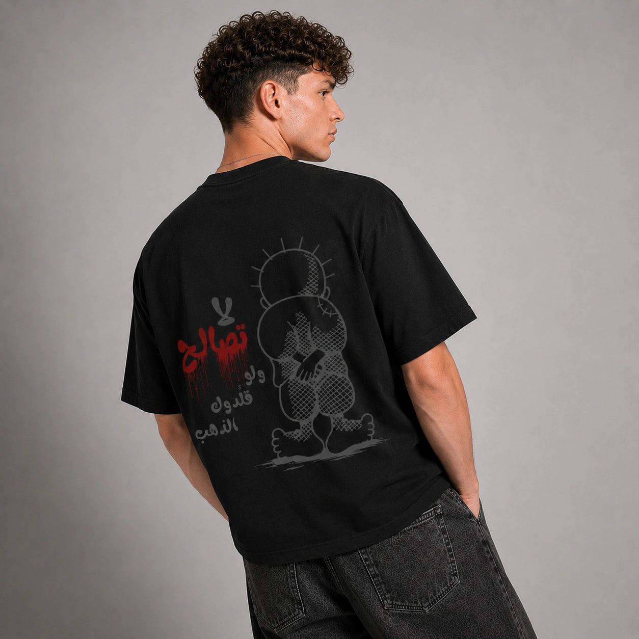 A man wearing a black graphic t-shirt, viewed from behind to show the back print. The shirt features a stylized cartoon character in gray and red Arabic text.