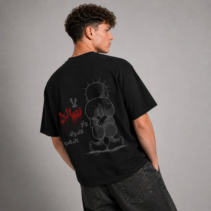 A man wearing a black graphic t-shirt, viewed from behind to show the back print. The shirt features a stylized cartoon character in gray and red Arabic text.