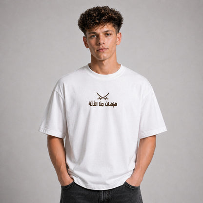 A young man with curly brown hair wearing a white crewneck t-shirt with a black graphic of crossed swords and Arabic text on the chest, posed against a neutral gray background.