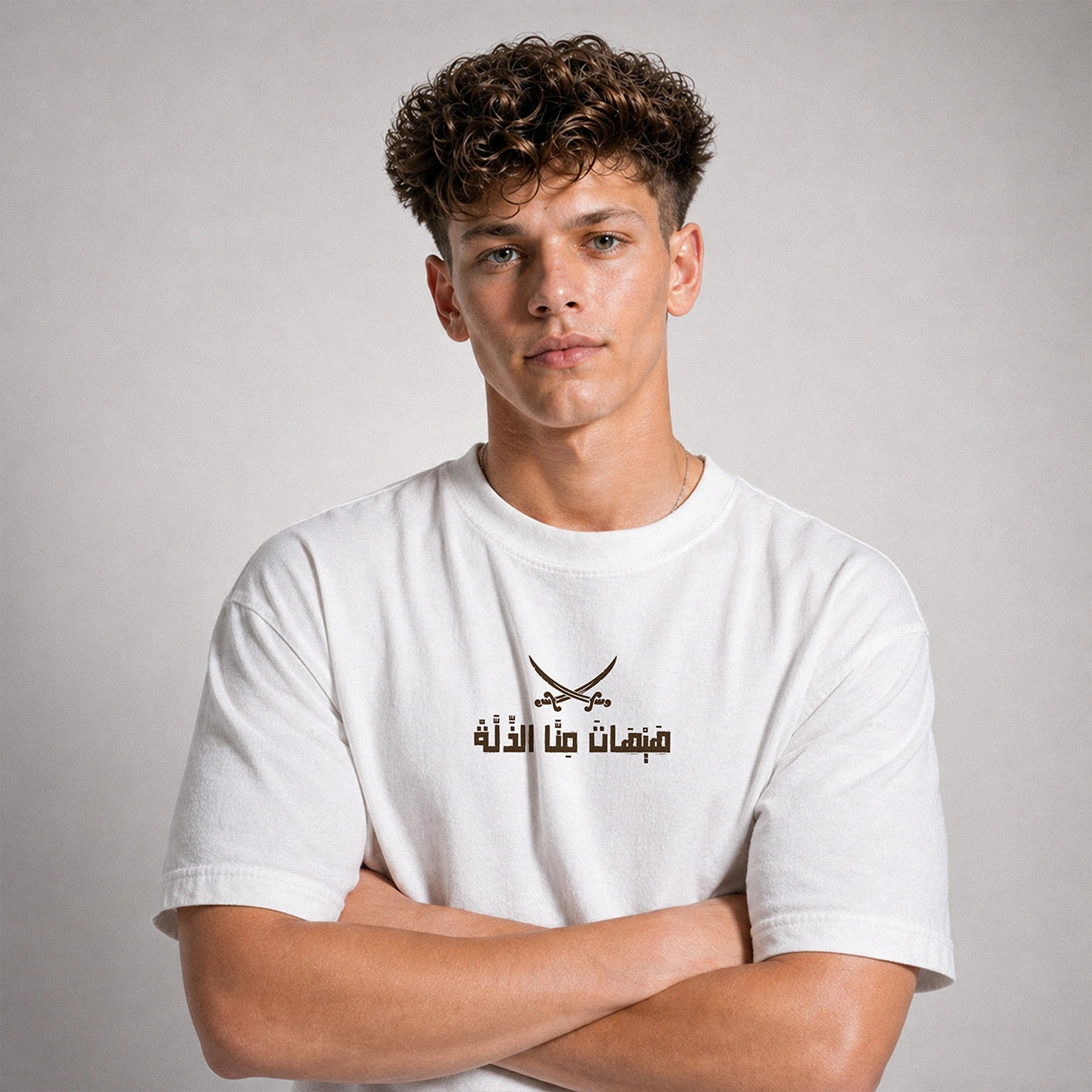 A young man with curly brown hair wearing a white short-sleeve t-shirt with a black printed graphic of crossed swords and Arabic text on the chest, posed with arms crossed against a neutral gray background.