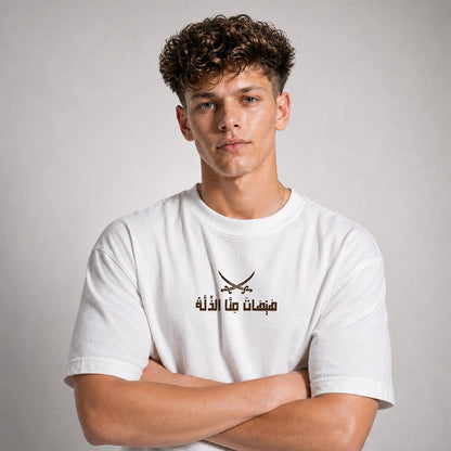 A young man with curly brown hair wearing a white short-sleeve t-shirt with a black printed graphic of crossed swords and Arabic text on the chest, posed with arms crossed against a neutral gray background.
