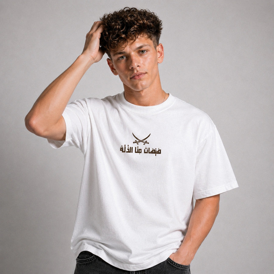 A young man with curly brown hair wearing a white crewneck t-shirt with a black graphic of crossed swords and Arabic text on the chest, posed against a neutral gray background.