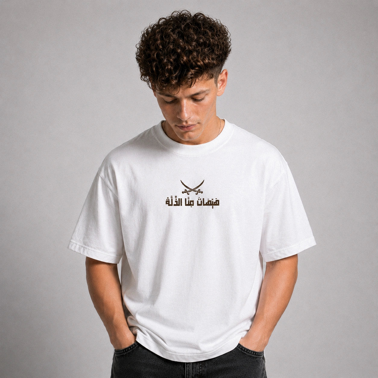 A young man with curly brown hair wearing a white crewneck t-shirt with a black graphic of crossed swords and Arabic text on the chest, posed against a neutral gray background.