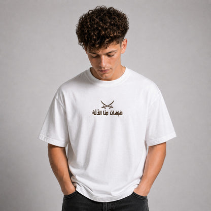 A young man with curly brown hair wearing a white crewneck t-shirt with a black graphic of crossed swords and Arabic text on the chest, posed against a neutral gray background.