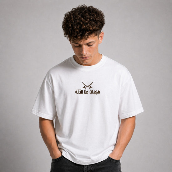 A young man with curly brown hair wearing a white crewneck t-shirt with a black graphic of crossed swords and Arabic text on the chest, posed against a neutral gray background.
