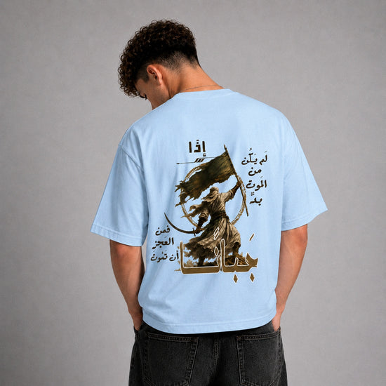 A man viewed from behind wearing a light blue t-shirt with a large graphic print on the back. The print features a dark brown illustration of a warrior with a sword and shield, surrounded by Arabic calligraphy.
