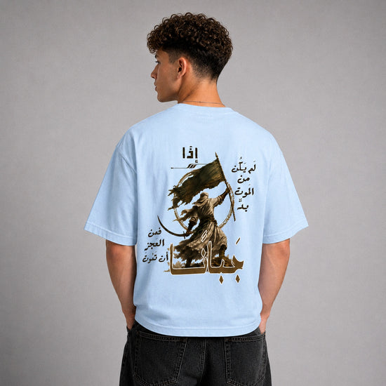 A man shown from the back wearing a light blue t-shirt with a large graphic print on the back. The graphic features a dark brown illustration of a figure holding a sword and flag, surrounded by Arabic text.