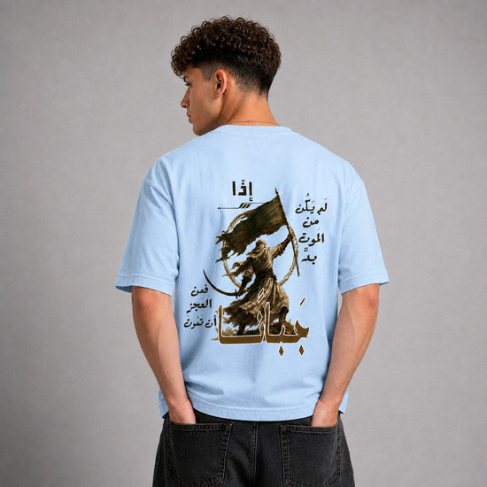 A man viewed from the back wearing a light blue t-shirt with a large graphic print on the back. The graphic features a dark brown silhouette of a figure holding a sword and a green flag, surrounded by Arabic calligraphy.