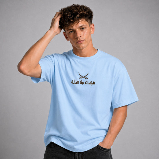 A young man with curly brown hair wearing a light blue crewneck t-shirt with a black graphic of crossed swords and Arabic text on the chest, posed against a neutral gray background.