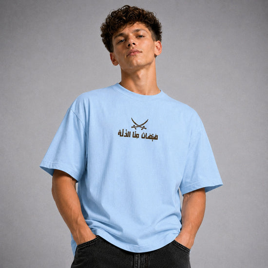 A young man with curly brown hair wearing a light blue crewneck t-shirt with a black graphic of crossed swords and Arabic text on the chest, paired with black jeans, set against a plain gray studio background.