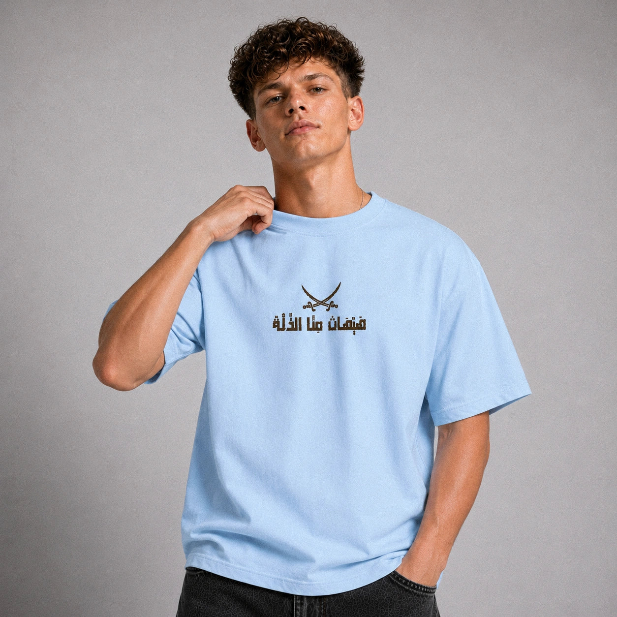 A young man wearing a light blue t-shirt with a black graphic of crossed swords and Arabic text on the chest, posed against a neutral gray studio background.