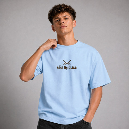 A young man wearing a light blue t-shirt with a black graphic of crossed swords and Arabic text on the chest, posed against a neutral gray studio background.