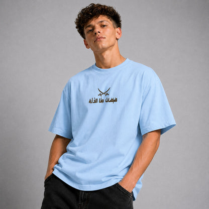 A young man wearing a light blue crewneck t-shirt with a black graphic of crossed swords and Arabic text on the chest, posed against a neutral gray background.