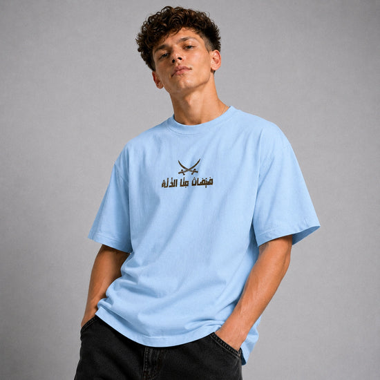 A young man wearing a light blue crewneck t-shirt with a black graphic of crossed swords and Arabic text on the chest, posed against a neutral gray background.