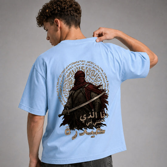 A man wearing a light blue t-shirt, viewed from the back. The shirt features a large graphic print of a hooded warrior holding a sword, surrounded by a circular gold pattern and Arabic calligraphy.