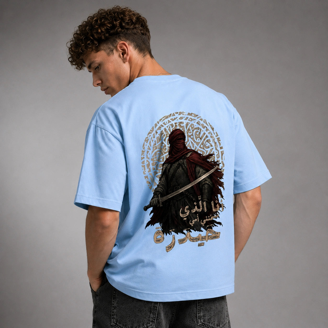 A man wearing a light blue t-shirt, shown from the back. The shirt features a large graphic print of a hooded figure holding a sword, surrounded by Arabic calligraphy in gold and red.