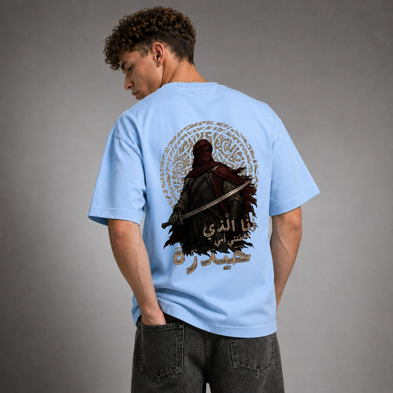 A man wearing a light blue t-shirt with a large graphic print on the back, shown from behind against a gray studio background.