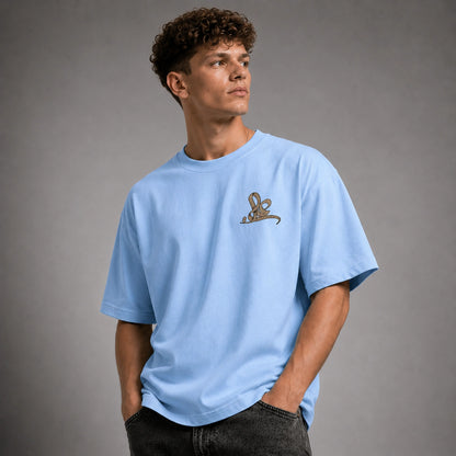 A man wearing a light blue oversized crewneck t-shirt with a small brown rabbit graphic on the left chest, paired with dark gray jeans, set against a solid gray studio background.