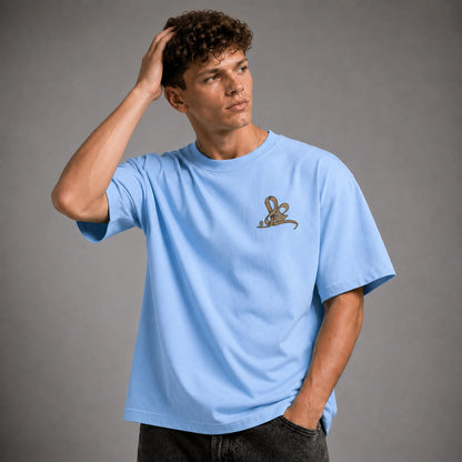 A man wearing a light blue crewneck t-shirt with a small brown embroidered graphic on the left chest, posed against a solid gray studio background.