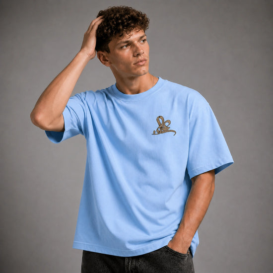 A man wearing a light blue crewneck t-shirt with a small brown embroidered graphic on the left chest, posed against a solid gray studio background.