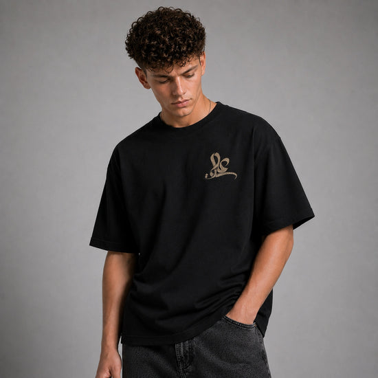 A man wearing a black oversized t-shirt with a small gold graphic logo on the left chest, paired with dark gray jeans, set against a plain gray studio background.
