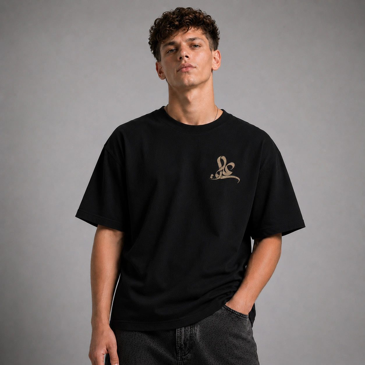 A man wearing a black short-sleeve t-shirt with a small gold script logo on the left chest, paired with dark gray jeans, photographed against a neutral gray studio background.