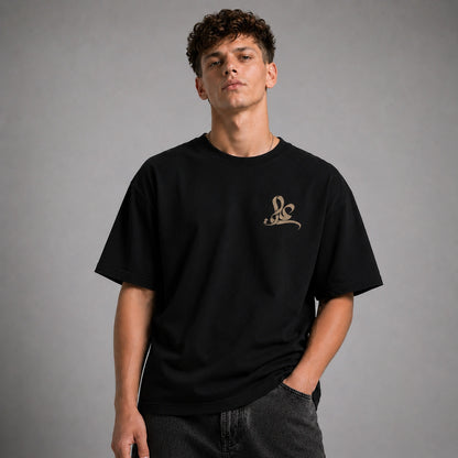 A man wearing a black short-sleeve t-shirt with a small gold script logo on the left chest, paired with dark gray jeans, photographed against a neutral gray studio background.