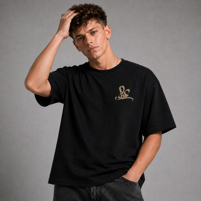 A man wearing a black short-sleeve t-shirt with a small metallic gold script logo on the left chest, posed against a neutral gray studio background.