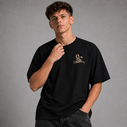 A young man with curly brown hair wearing a black short-sleeve t-shirt with a small gold graphic logo on the left chest, paired with dark gray jeans, set against a solid gray studio background.