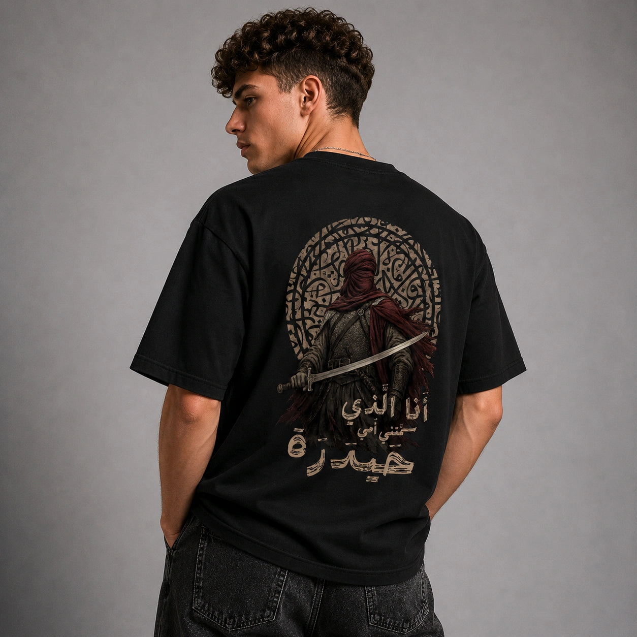 A man wearing a black t-shirt with a large graphic print on the back, shown from behind against a gray studio background.