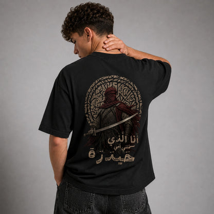 A man wearing a black t-shirt with a large graphic print on the back, shown from behind against a gray studio background.