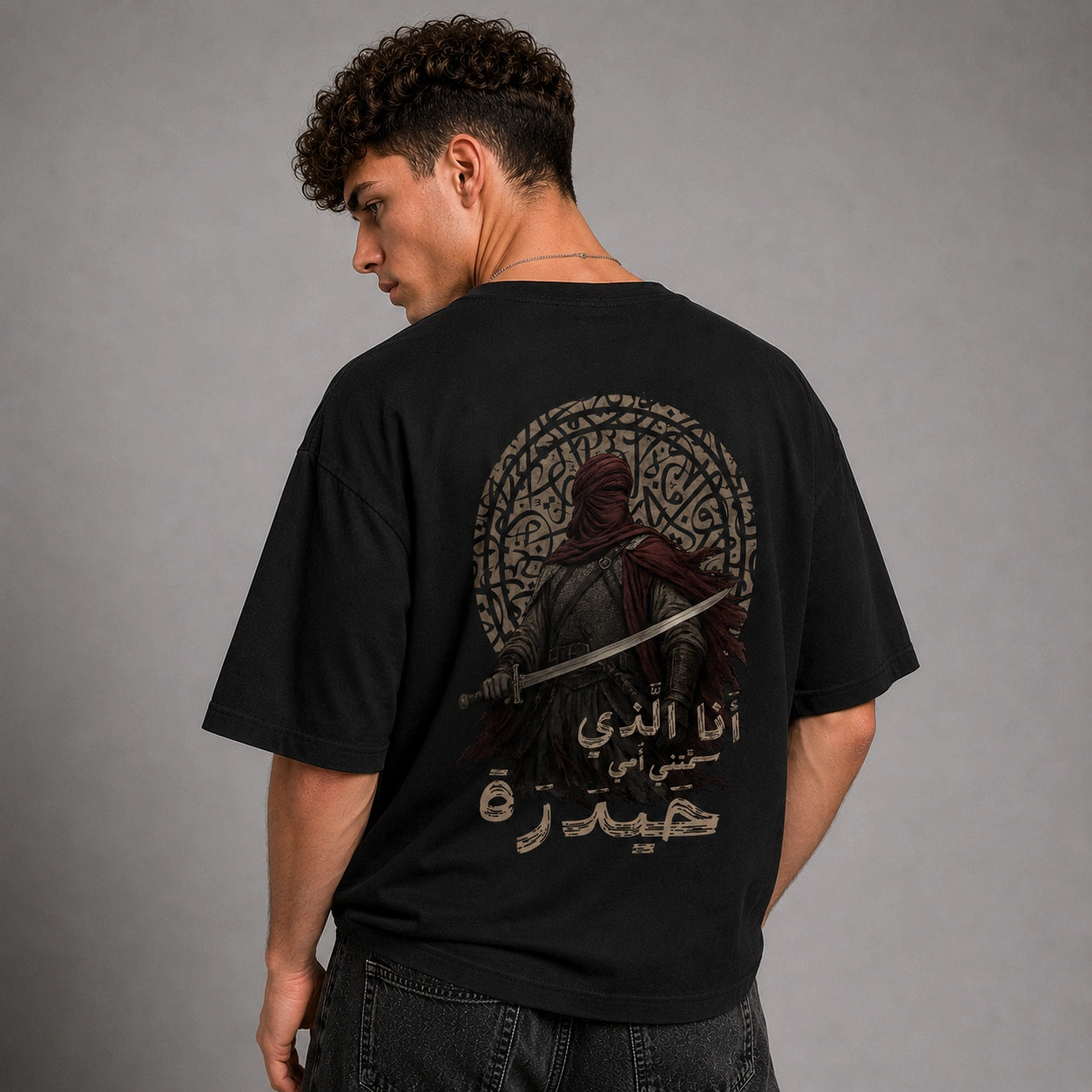 A man wearing a black graphic t-shirt, viewed from the back. The shirt features a large circular design with a hooded figure holding a sword, surrounded by intricate Arabic calligraphy.