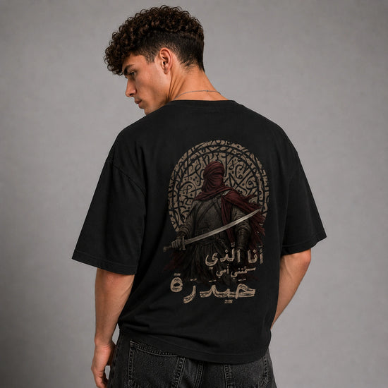 A man wearing a black graphic t-shirt, viewed from the back. The shirt features a large circular design with a hooded figure holding a sword, surrounded by intricate Arabic calligraphy.