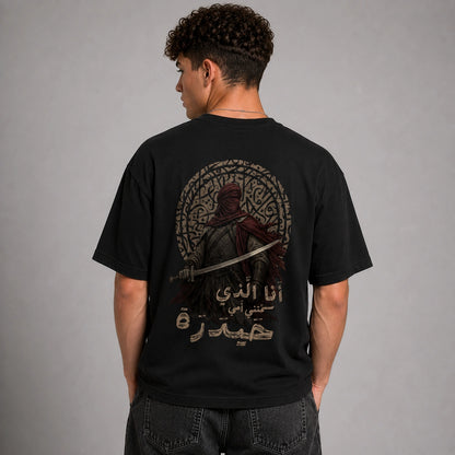 A man wearing a black t-shirt from the back, showing a large graphic print of a hooded warrior with a sword inside a circular frame, with Arabic text below. The model is standing against a neutral gray studio background.