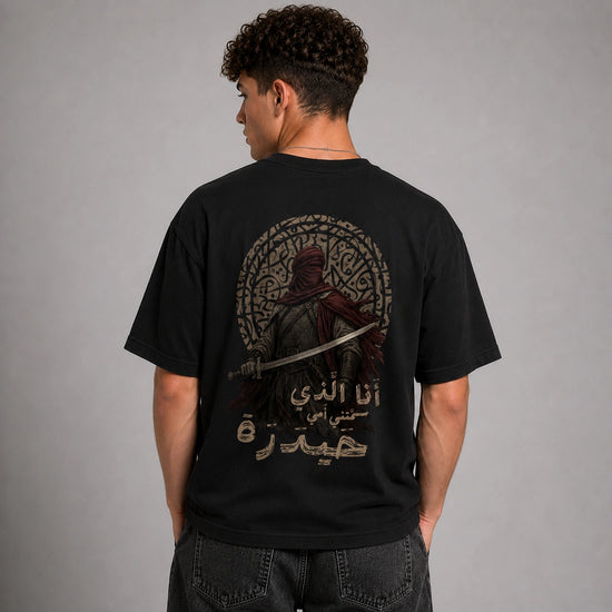A man wearing a black t-shirt from the back, showing a large graphic print of a hooded warrior with a sword inside a circular frame, with Arabic text below. The model is standing against a neutral gray studio background.