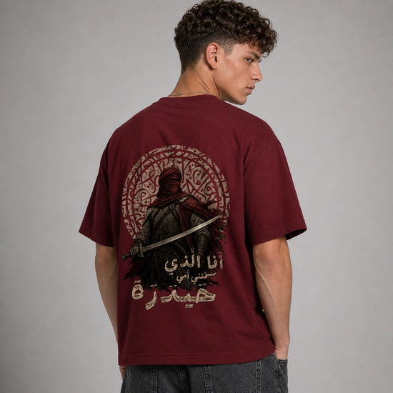 A man wearing a maroon t-shirt from the back, featuring a large graphic print of a hooded warrior holding a sword inside a circular patterned frame with Arabic text.