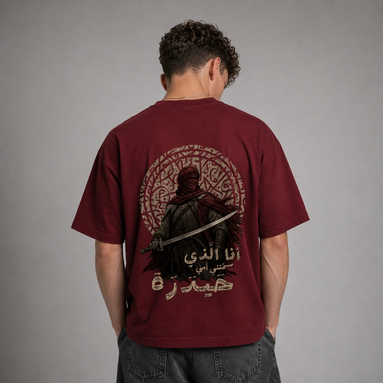 A man viewed from behind wearing a maroon short-sleeve t-shirt with a large graphic print on the back. The graphic features a dark figure holding a sword, an intricate circular pattern, and Arabic text.