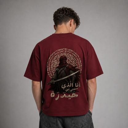 A man viewed from behind wearing a maroon short-sleeve t-shirt with a large graphic print on the back. The graphic features a dark figure holding a sword, an intricate circular pattern, and Arabic text.