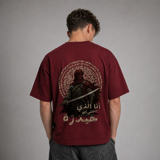 A man viewed from behind wearing a maroon short-sleeve t-shirt with a large graphic print on the back. The graphic features a dark figure holding a sword, an intricate circular pattern, and Arabic text.