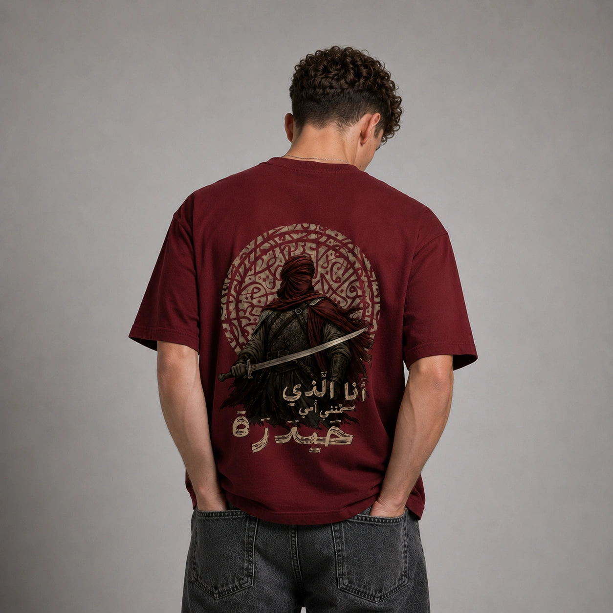 A man viewed from behind wearing a maroon short-sleeve t-shirt with a large graphic print on the back, paired with dark gray jeans, against a neutral gray studio background.