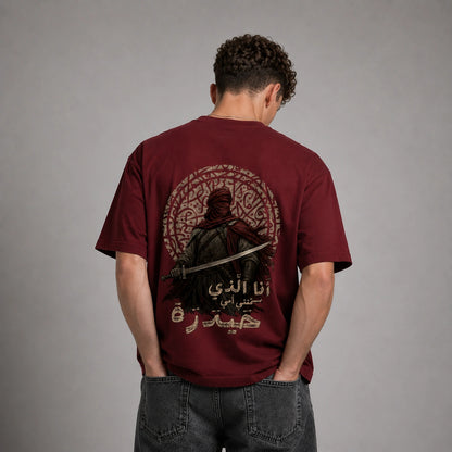 A man viewed from behind wearing a maroon short-sleeve t-shirt with a large graphic print on the back, paired with dark gray jeans, against a neutral gray studio background.
