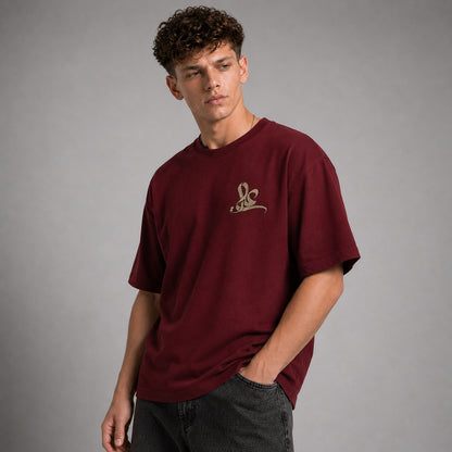 A young man wearing a maroon short-sleeve t-shirt with a small gold embroidered logo on the left chest, paired with dark gray jeans, photographed against a neutral gray studio background.