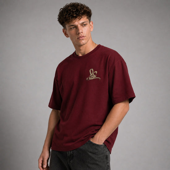 A young man wearing a maroon short-sleeve t-shirt with a small gold embroidered logo on the left chest, paired with dark gray jeans, photographed against a neutral gray studio background.